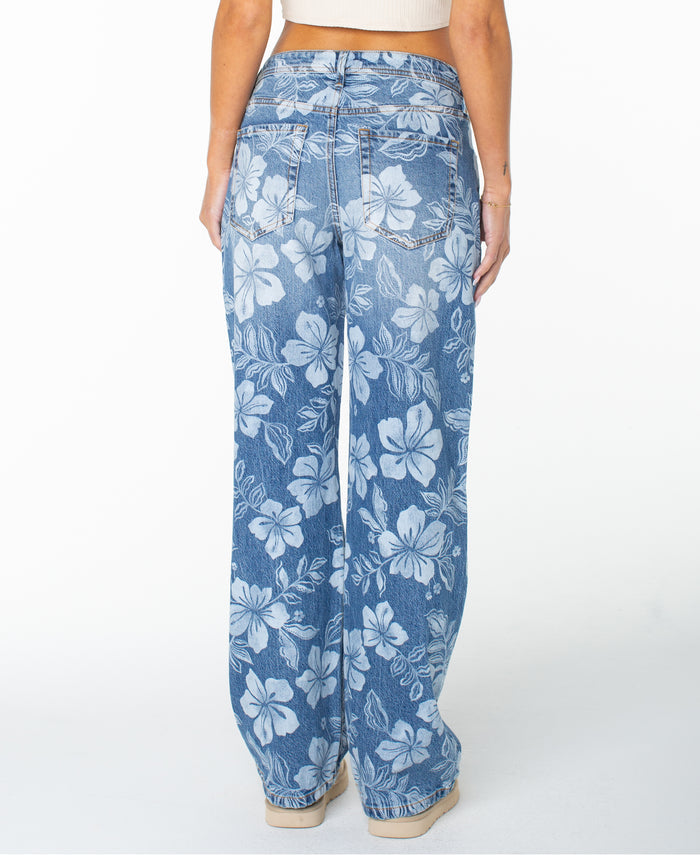 Model showcases the Roxy Haven Flora Pant, styled in a relaxed oversized fit with a wide-leg silhouette and bold white hibiscus floral print. Seen from the back, these pants feature two back pockets and tan stitching, effortlessly paired with beige shoes and a light crop top.