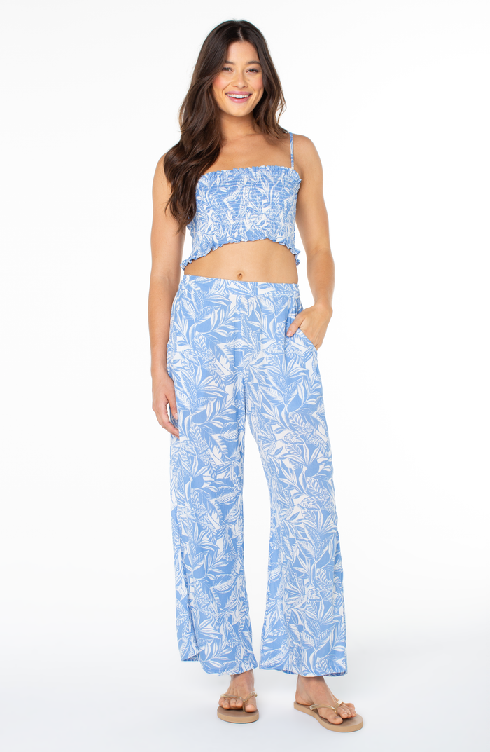 A woman smiles with one hand in her pocket, wearing the Roxy Hello Sunshine Top in light blue floral and matching wide-leg pants, paired with tan sandals against a white background.