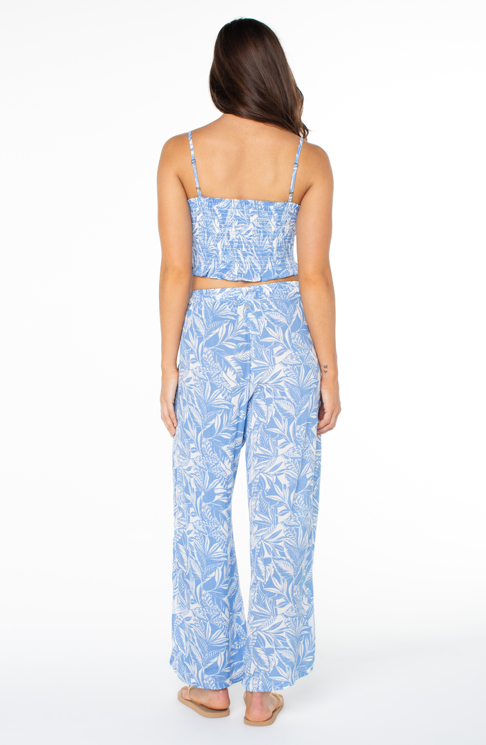 A woman with long brown hair stands barefoot, facing away, wearing the Roxy Hello Sunshine Top in light blue with a white leafy pattern and matching wide-leg pants. The background is plain white.