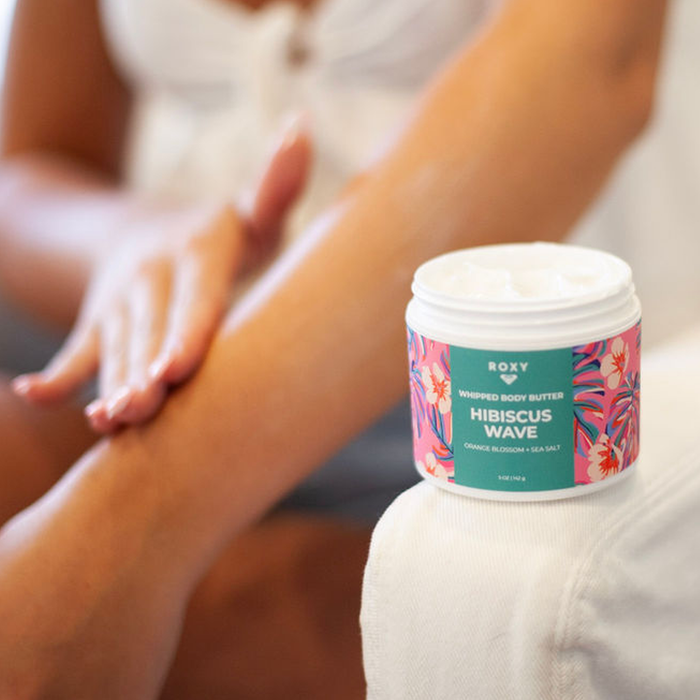 A person applies lotion to their leg while a jar of Roxy Hibiscus Wave Body Butter, with a colorful floral label, sits on a white surface—ideal for daily hydration and soothing dry skin.