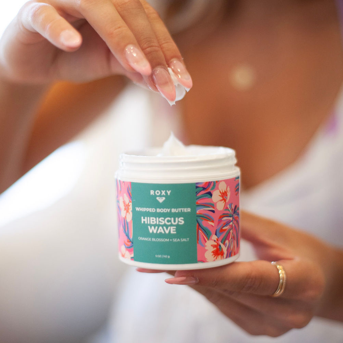 A woman with manicured nails and a gold ring scoops Roxy Hibiscus Wave Body Butter from a vibrant jar, offering daily hydration and relief for dry skin.