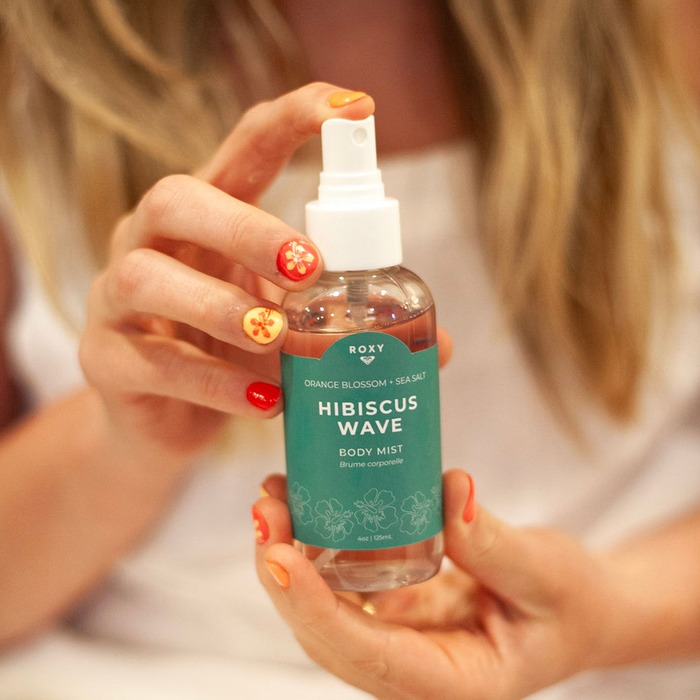 A person with long blonde hair and floral nail art holds a Roxy Hibiscus Wave Body Mist, featuring a teal label and white spray nozzle, evoking fresh coconut and citrus notes.