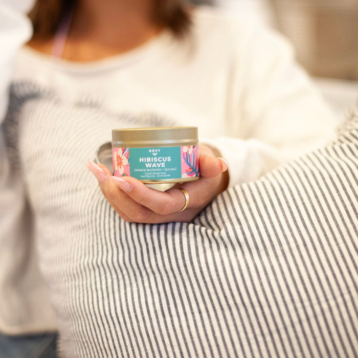 A person in a white shirt holds the Roxy Hibiscus Wave Palm Tin Candle with a vibrant floral design, suggesting tropical scents, while their hand rests on a striped pillow. Their face is out of focus.