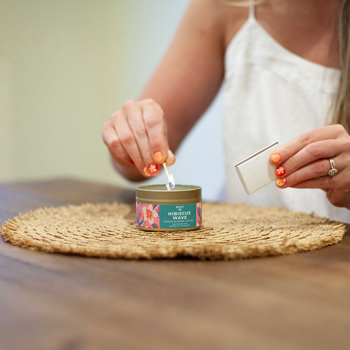 A woman with orange floral nail art lights a Roxy Hibiscus Wave Palm Tin Candle on a woven placemat, filling the air with tropical scent. She wears a white top and an elegant ring on her left hand.