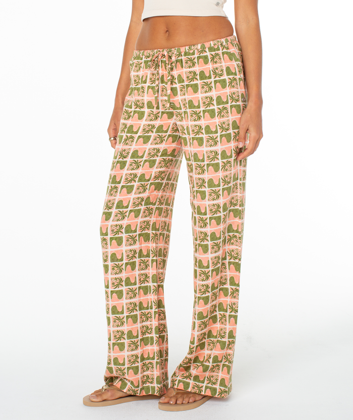 A person in the Roxy Iyla Pant 2, featuring a pink and green geometric floral pattern and relaxed low-rise fit, styled with a beige crop top and sandals, stands against a white background for an effortlessly coastal vibe.