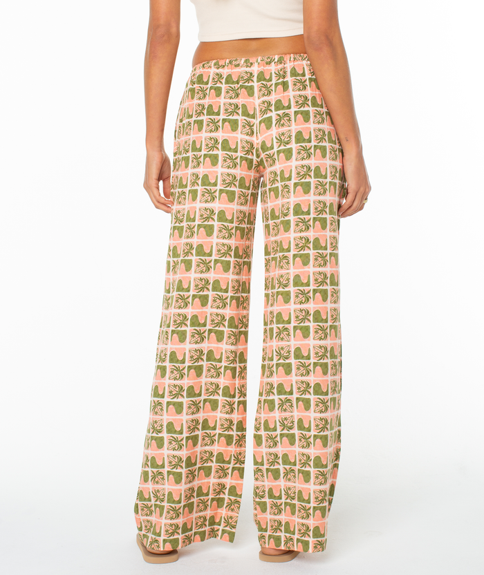 A person models the Roxy Iyla Pant 2 in a green and peach retro geometric print, facing away to show the back. The relaxed low-rise fit pairs with a white crop top and sandals for easy coastal style.