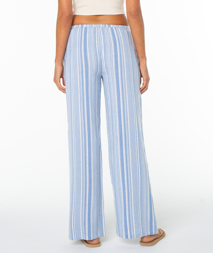 A person stands facing away against a white background, wearing the Roxy Iyla Pant 2 in light blue and white vertical stripes with a relaxed fit, styled with a white crop top and sandals for an effortless coastal look.