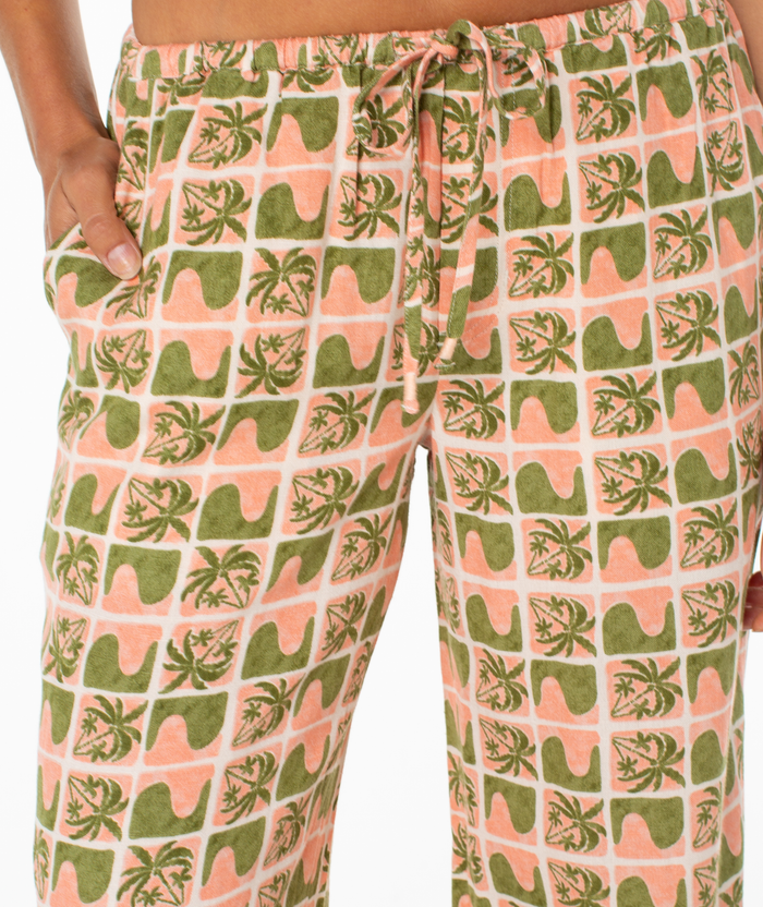 Close-up of Roxy’s Iyla Pant 2 for women—a relaxed, low-rise peach pant with olive-green tropical palm, surfboard, and wave print in a grid pattern. Side pocket adds effortless coastal charm.