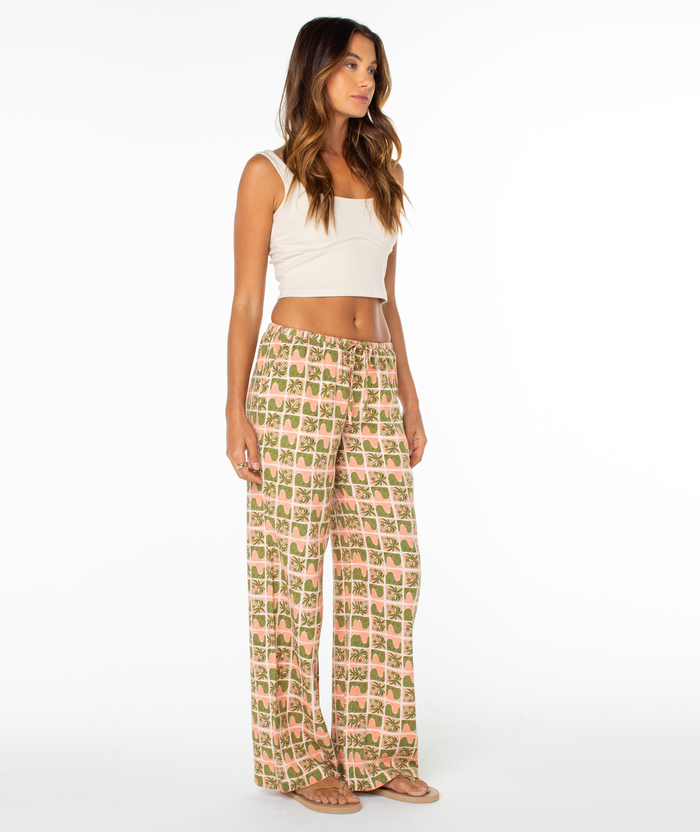 A woman stands barefoot against a white background, wearing a white crop tank and Roxy's Iyla Pant 2, which features a relaxed low-rise fit with a green and pink square pattern. She looks slightly to the side, radiating coastal charm.