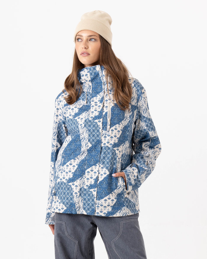 A person wearing a beige beanie, gray pants, and the Roxy Jetty 3N1 Jacket in Loom State—a blue and white patterned women’s snowboard jacket made from recycled polyester—stands against a plain white background with one hand in their pocket, looking to the side.