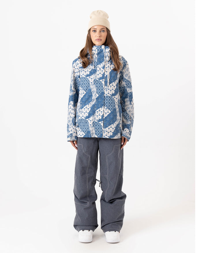 A person stands facing forward against a plain white background wearing a beige beanie, loose gray pants, white shoes, and the Roxy Jetty 3N1 Jacket—a blue and white patterned women’s snowboard jacket made from recycled polyester.