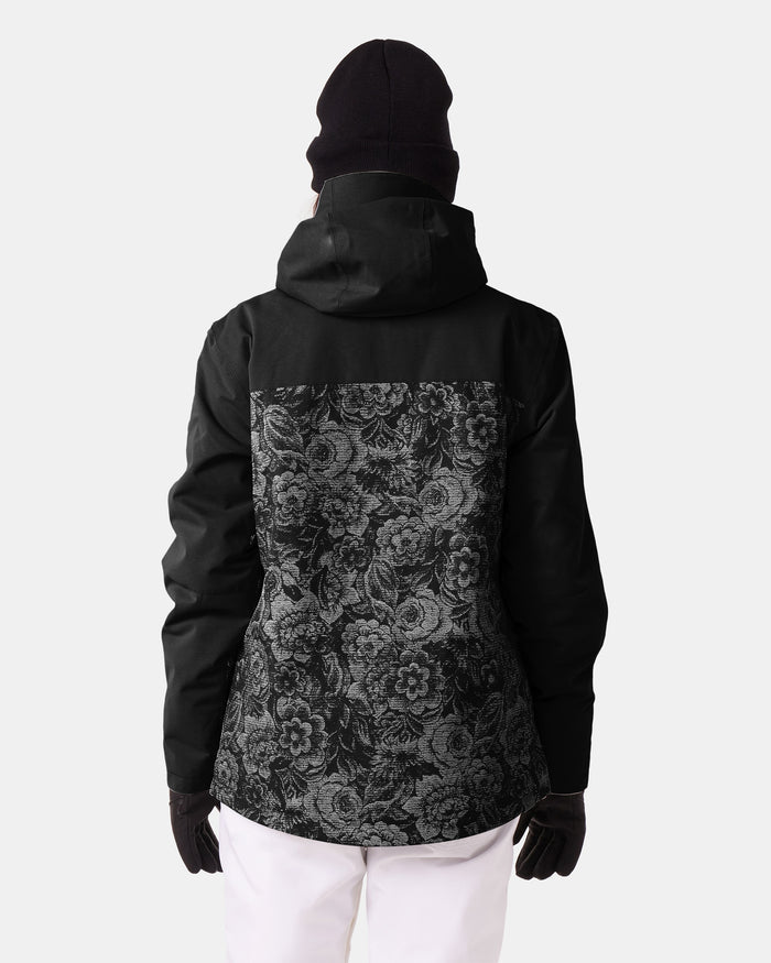 A person wearing a black beanie and gloves is shown from behind against a plain light background. They are wearing the Roxy Jetty 3N1 Jacket—a women’s snowboard jacket with a 3-in-1 design, featuring a black exterior with a floral pattern on the back—paired with white pants.
