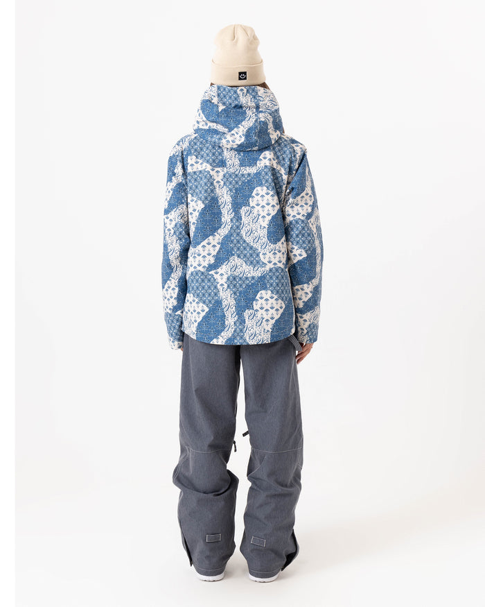 A person stands facing away, wearing the Roxy Jetty 3N1 Jacket—an eco-friendly women’s snowboard jacket in a blue and white pattern made from recycled polyester—paired with gray baggy pants and a beige beanie, set against a plain white background.