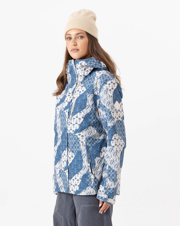 A woman wearing the Roxy Jetty 3N1 Jacket, featuring a blue and white pattern and made from recycled polyester, paired with gray pants and a cream beanie, stands against a plain white background looking slightly to the side.
