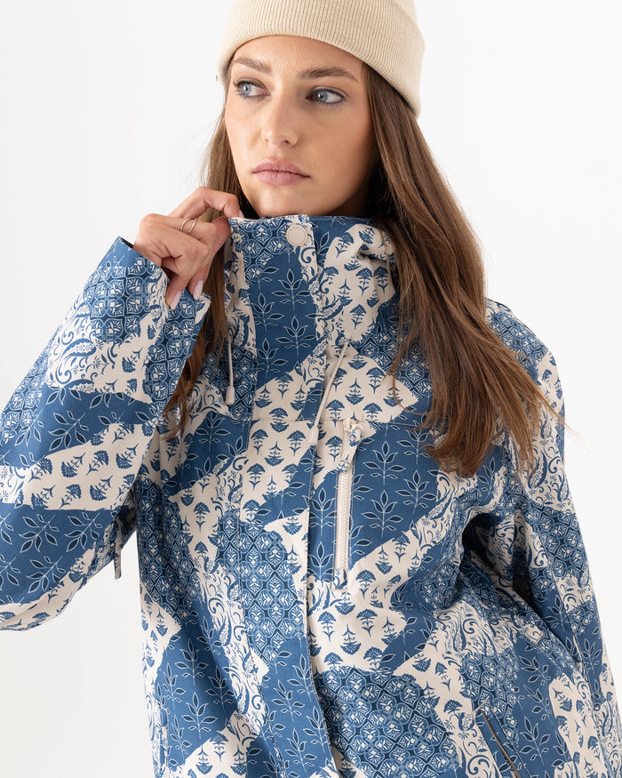 A woman wears the Roxy Jetty 3N1 Jacket, a blue-and-white patterned women’s snowboard jacket, paired with a beige beanie. She looks to the side and lifts the collar with one hand against a plain white background. Made from recycled polyester, this 3-in-1 jacket combines style and sustainability for all your winter adventures.
