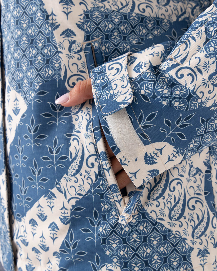 A hand with light purple nail polish is slipping into the side pocket of a blue and white patterned Roxy Jetty 3N1 Jacket for women, featuring floral and leaf designs crafted from recycled polyester.