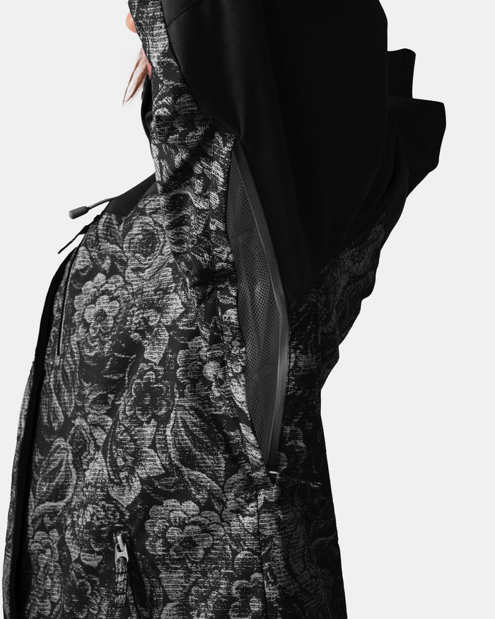 A woman wearing the Roxy Jetty 3N1 Jacket in a black and gray floral pattern raises her arm, revealing a zippered mesh underarm vent that adds comfort. The waterproof and breathable design is displayed against a plain white background.