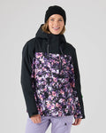 A person wearing a black beanie and the Roxy Jetty Jacket in Blended Bouquet—a women’s snowboard jacket with a floral pattern in shades of purple, pink, and black—stands against a plain light background, smiling at the camera. The Jetty Jacket features warm insulation and is made from recycled materials for comfort and sustainability.