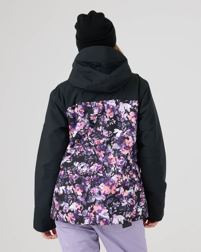 A person is seen from behind against a plain light background, wearing the Roxy Jetty Jacket—a black women’s snowboard jacket with a floral purple and pink back panel, warm insulation, and crafted from recycled materials—paired with a black beanie and light purple pants.