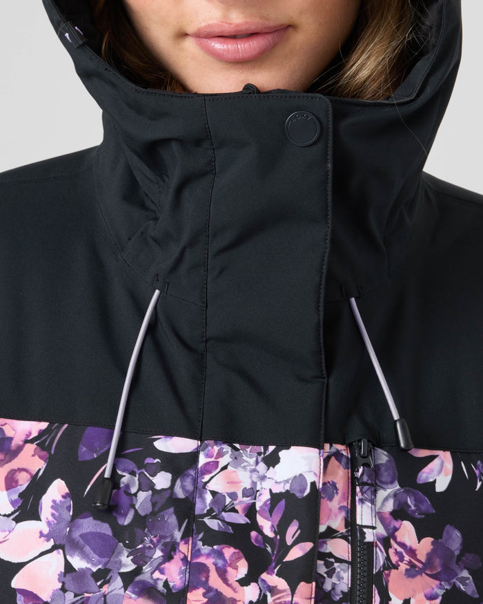 A close-up of a woman wearing the Roxy Jetty Jacket, a black women’s snowboard jacket featuring purple floral patterns and white drawstrings, made from recycled materials. Only the lower half of her face is visible.