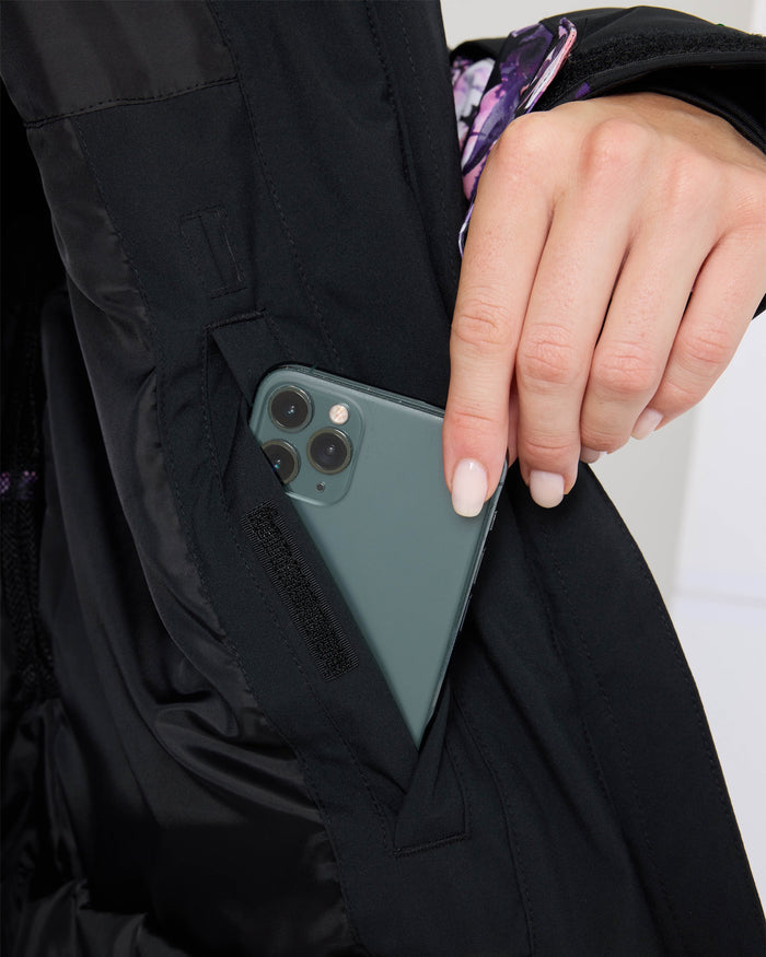 A hand with manicured nails places a green smartphone into the inner pocket of the Roxy Jetty Jacket, a black women’s snowboard jacket crafted with warm insulation and recycled materials.