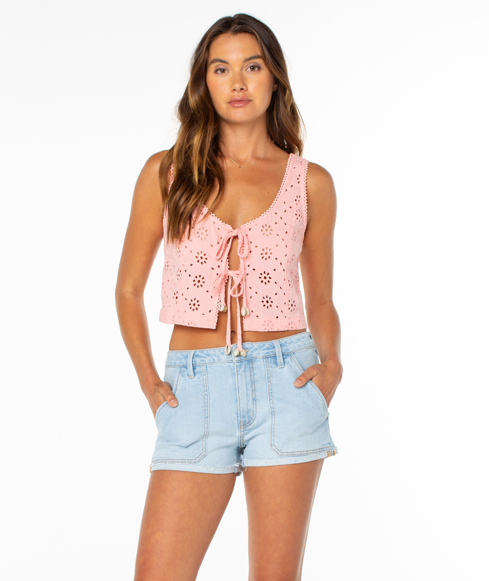 A woman with long brown hair wears the Roxy Kayla Tank, a pink crochet eyelet top with front ties and a scoop neckline, paired with light blue denim shorts. She stands with hands in her pockets against a plain white background.