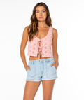 A woman with long brown hair wears the Roxy Kayla Tank, a pink crochet eyelet top with front ties and a scoop neckline, paired with light blue denim shorts. She stands with hands in her pockets against a plain white background.