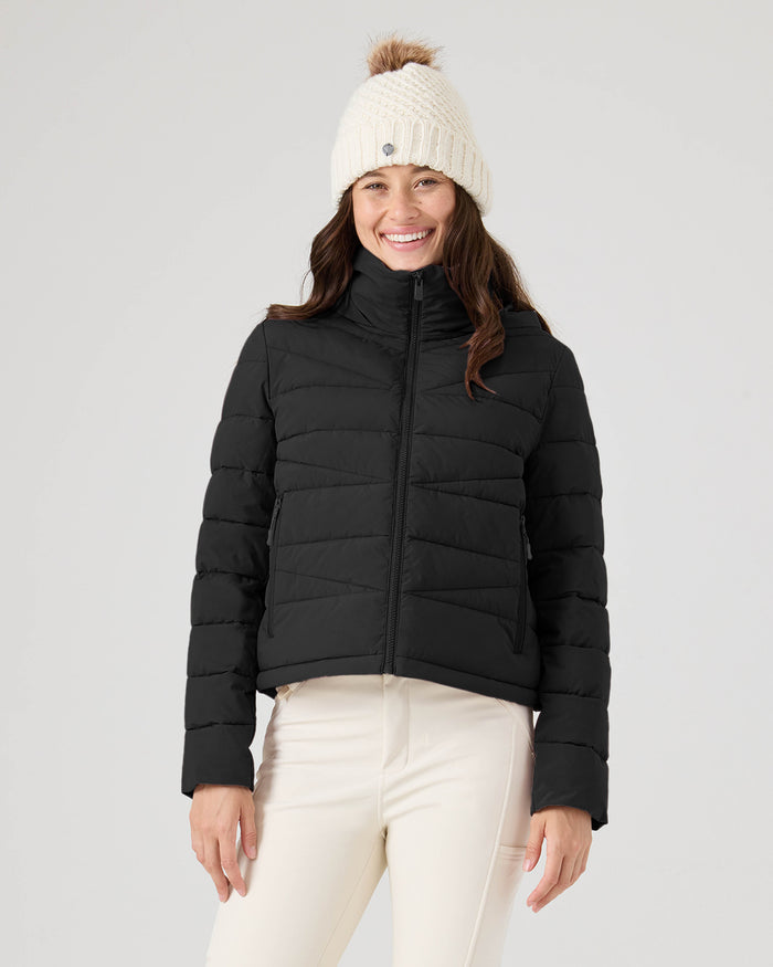 A woman stands smiling against a plain light background, wearing a cream knit pom-pom beanie, white pants, and the Roxy Kelly Lightweight Puffer in black.