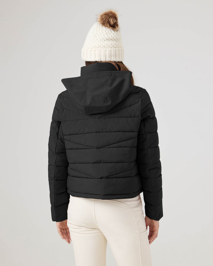 A person shown from the back against a plain light background is wearing the Roxy Kelly Lightweight Puffer in black, paired with a white knit beanie featuring a brown pom-pom and cream pants.