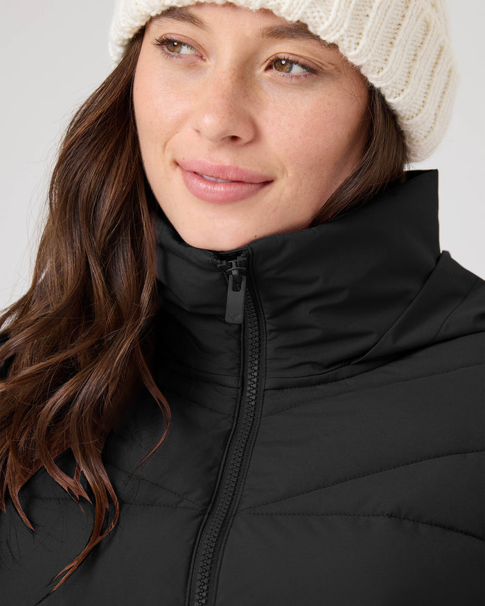 A person wearing a white knit beanie and the Roxy Kelly Lightweight Puffer in black looks slightly to the side, smiling softly.