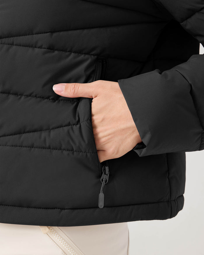 A close-up of a person wearing the Roxy Kelly Lightweight Puffer in black, unzipping a side pocket with their left hand. The person is also wearing light-colored pants.
