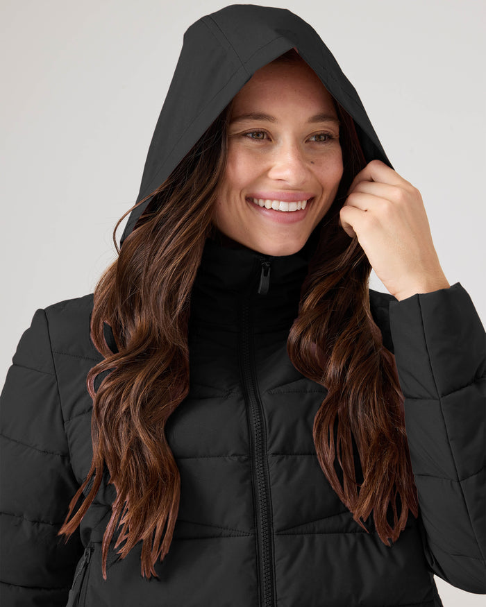 A person with long brown hair smiles while gently holding the hood of the black Kelly Lightweight Puffer by Roxy.