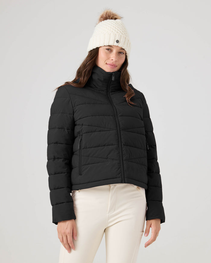 A woman wearing the Roxy Kelly Lightweight Puffer, paired with a white knit pom-pom hat and cream pants, stands against a plain light background, looking at the camera with a slight smile.