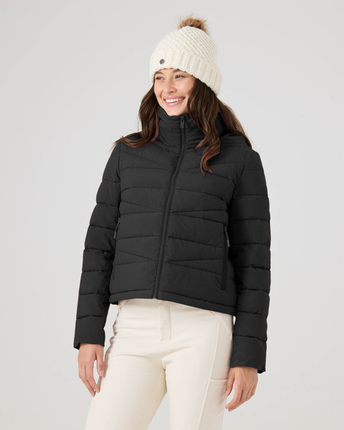 A woman wearing the Roxy Kelly Lightweight Puffer, paired with a cream knit beanie with a pom-pom and white pants, stands smiling against a plain light background.