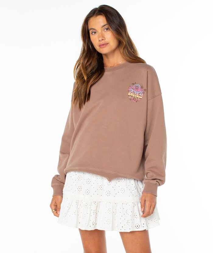 A woman with long brown hair wears the Roxy Lineup Oversized Crew sweatshirt, featuring a chest graphic, paired with a white eyelet ruffle skirt and stands against a plain white background.