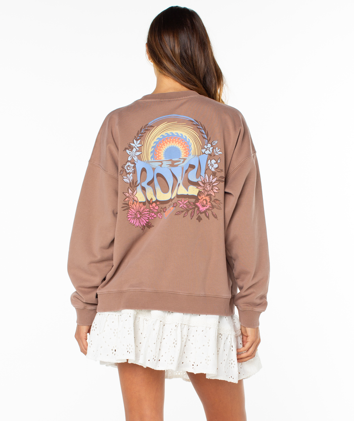A woman faces away wearing the Roxy Lineup Oversized Crew in brown with colorful flower and sunrise graphics and "ROXY," paired with a white, ruffled eyelet skirt against a plain white background.