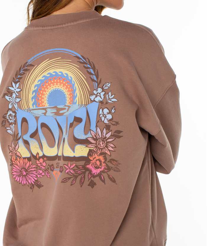 A person is shown from the torso up wearing Roxy's Lineup Oversized Crew, a mauve cotton blend sweatshirt with a colorful back graphic featuring "ROXY," flowers, and a retro sun and wave design.