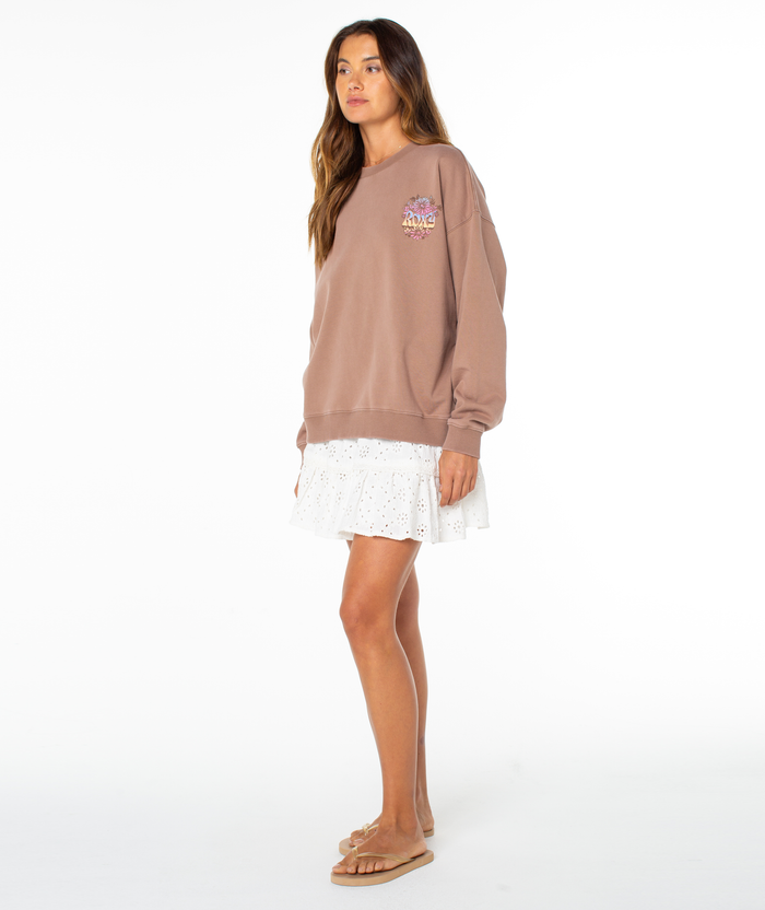 A woman with long brown hair and a neutral expression wears the Roxy Lineup Oversized Crew in brown, paired with a white eyelet skirt and beige slide sandals, standing sideways against a plain white background.