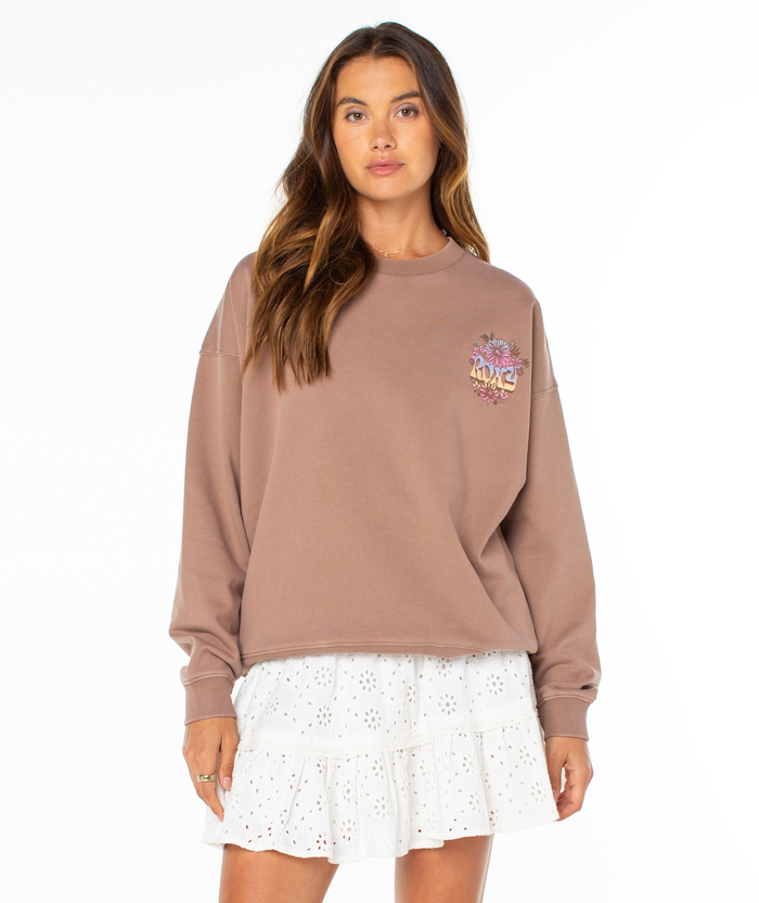A woman with long brown hair wears the Roxy Lineup Oversized Crew in a mauve sun-washed finish with a floral NYC graphic, paired with a white eyelet-embroidered tiered mini skirt, standing against a plain white background.