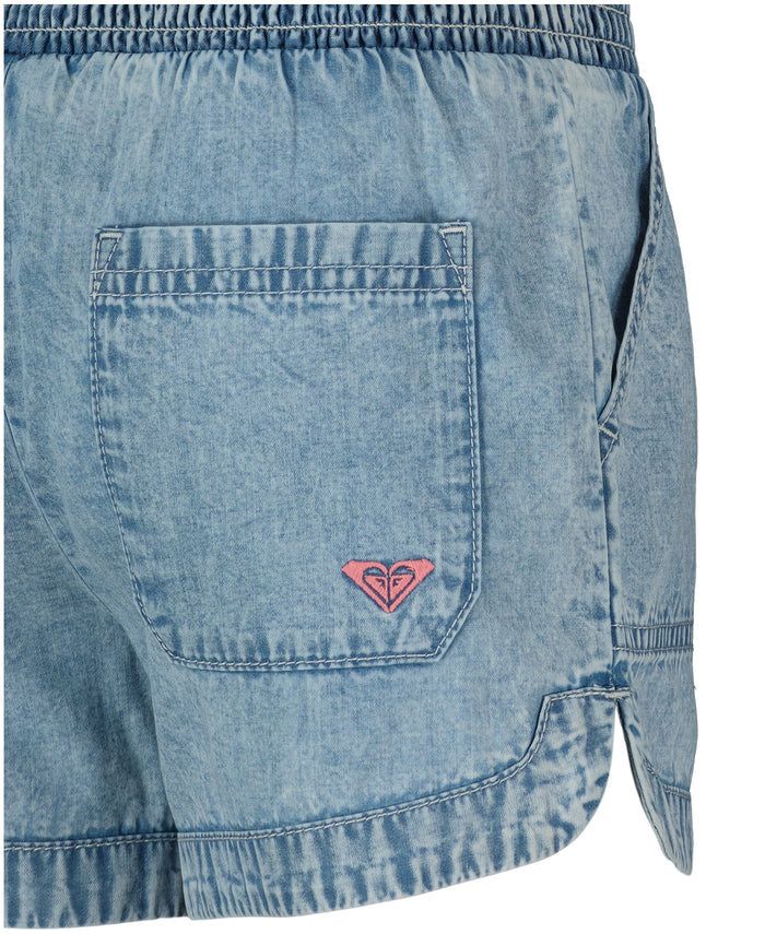 Close-up of the back and side of Roxy Little Girls' Soleil Short, featuring lightweight denim, a smocked elastic waistband, a back pocket with a small pink embroidered logo, and visible stitching details.