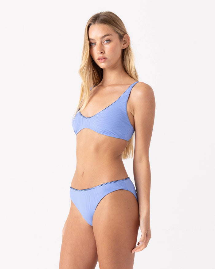 A woman with long blonde hair wears Roxy's Loop Me In Hipster Full Bikini Bottom in light blue with a ribbed texture, standing against a plain white background and gazing slightly to the side with a neutral expression.