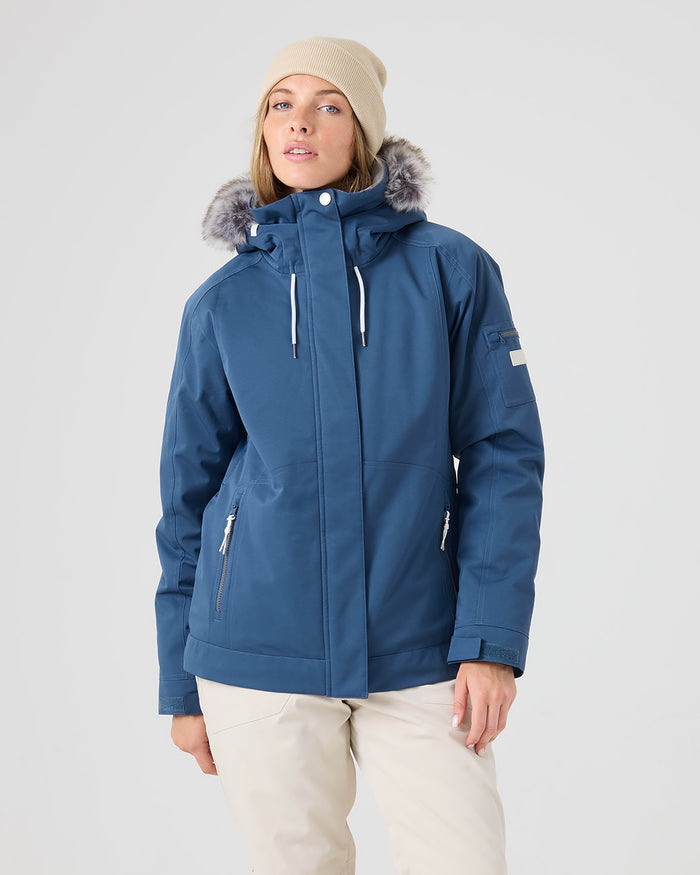 A person wearing a beige beanie, light pants, and a Roxy Meade Jacket in Ensign Blue with a faux fur-lined hood stands against a plain gray background.