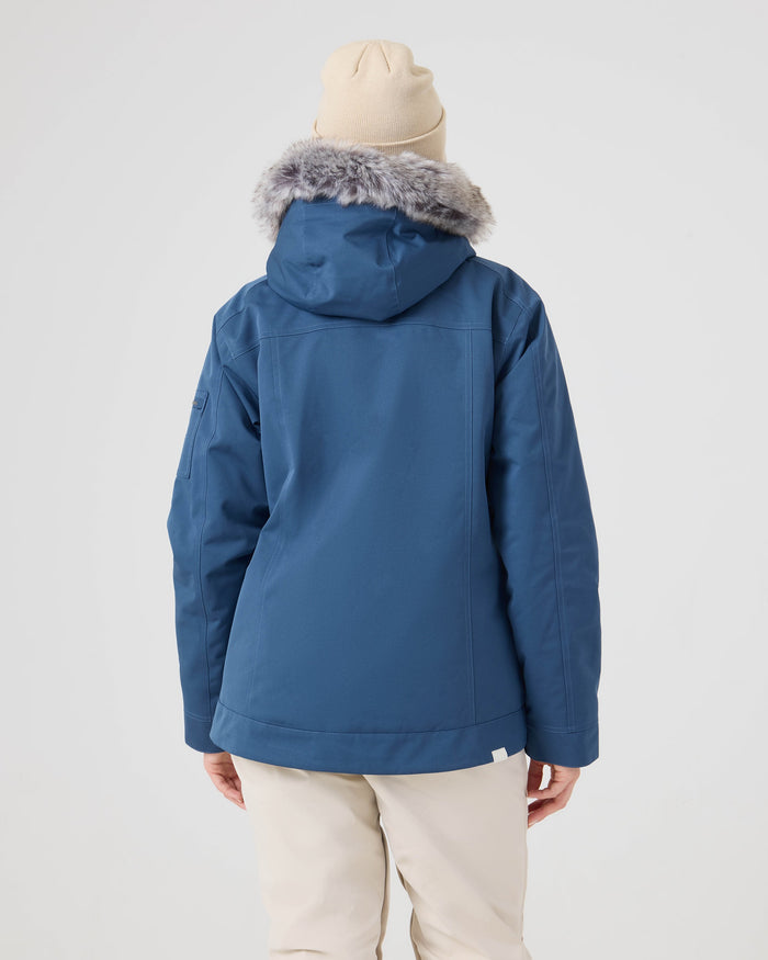 A person faces away from the camera wearing the Roxy Meade Jacket, a blue women’s snow jacket with a faux fur hood, paired with a beige beanie and light-colored pants against a plain background.