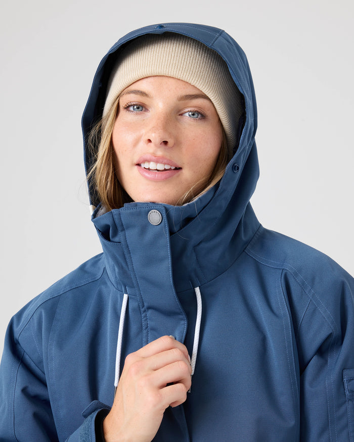 A person wearing the Roxy Meade Jacket, a blue hooded winter ski jacket, and a beige beanie looks at the camera, smiling slightly, with one hand holding the jacket collar.