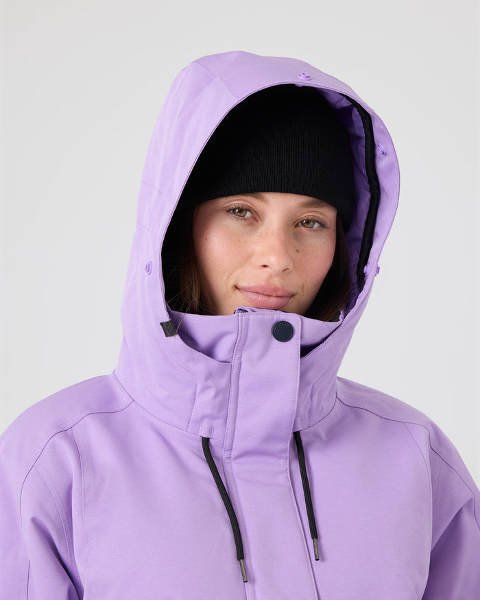 A person wearing a black beanie and the Roxy Meade Jacket—a lavender women’s snow jacket with a tailored fit—looks at the camera, smiling slightly. The jacket’s hood is up, offering both style and 10K protection, set against a plain light gray background.