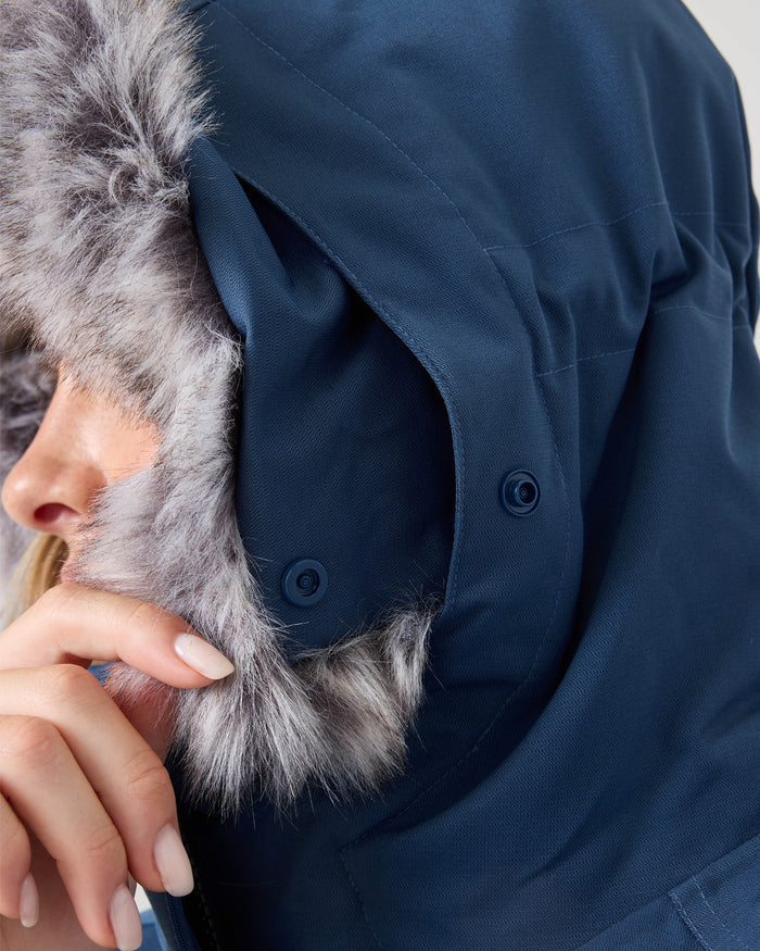 Close-up of a person wearing the Roxy Meade Jacket, a blue women’s snow jacket with a furry hood, holding the hood near their face with manicured fingers. The fur trim and fabric details of this winter ski jacket are clearly visible.