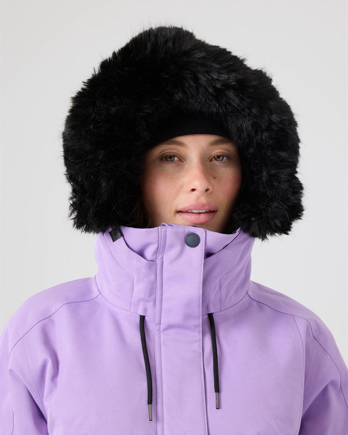 A person is wearing the Roxy Meade Jacket, a light purple women’s snow jacket with 10K protection, high collar, and a black fur-lined hood, standing against a plain light background.