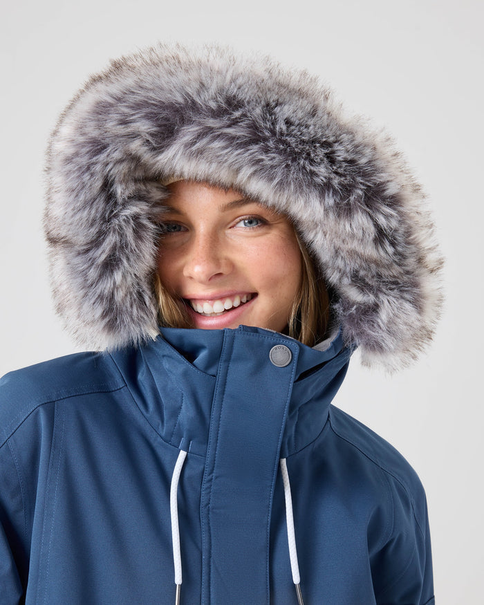 A person smiling, wearing the Roxy Meade Jacket—a blue women’s snow jacket featuring a hood trimmed in fluffy gray faux fur—against a plain light background.