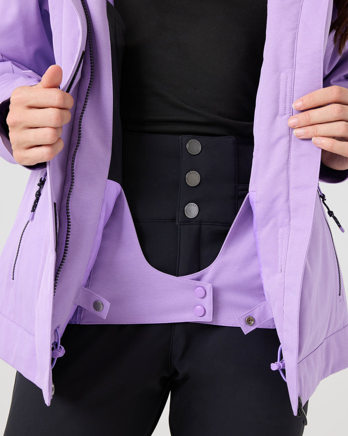 A person wears a black shirt, black pants with three buttons, and the Roxy Meade Jacket—a light purple women’s snow jacket with a tailored fit—partially unzipped to showcase an inner waistband adorned with purple snap buttons. With hands holding the jacket open, its chic design and dependable 10K protection are highlighted.