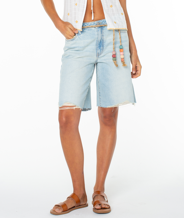 A person wearing Roxy's Message In A Bottle Short in light blue with frayed edges, paired with a white sleeveless top with rope tie and drawstring waist, plus brown sandals, stands against a white background.